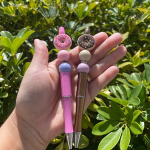 May include: Two pens with donut-shaped tops. One pen is pink with a pink and purple beaded section and a pink donut top with colorful sprinkles. The other pen is brown with a cream and pink beaded section and a brown donut top with white sprinkles.