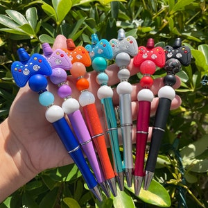 May include: A set of eight colorful pens with a video game controller design. The pens are decorated with beads and have a silver metal tip.