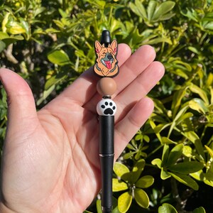 May include: A black pen with a brown and white paw print charm and a brown and tan German Shepherd charm.