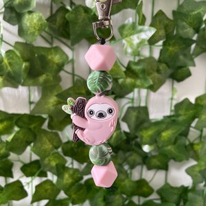May include: A pink and green sloth keychain with a silver clip. The sloth is hanging from a branch and has a pink body and a brown branch. The keychain has a pink, green, and white bead pattern.