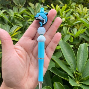 May include: A turquoise pen with a dolphin-shaped topper. The dolphin is black and blue with a smiling face. The pen has a bamboo-like design and silver accents. The pen is held against a background of green leaves.