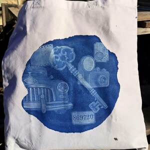 May include: A white canvas tote bag with a blue design. The design features a vintage car, a key, and various cameras. The number 869720 is printed on the bag.