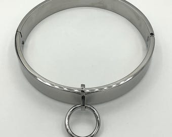 May include: A polished silver-colored metal collar with a hinged design and a small ring pendant. The collar is circular and has a smooth, reflective surface. The ring pendant hangs from the center of the collar.