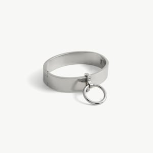 May include: A polished silver-tone metal hinged bangle bracelet featuring a smooth, wide band design. A circular ring charm is attached to the front of the band by a small vertical metal connector, creating a clean and reflective metallic aesthetic.