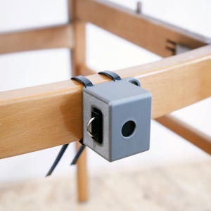Op de afbeelding: Een grijs kubusvormig meubelaccessoire is met zwarte bandjes aan een houten stoelpoten bevestigd. De kubus heeft een rond gat en een metalen ring. De stoelpoten zijn lichtbruin.