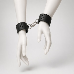 May include: A pair of matte black wrist cuffs connected by a silver-toned metal carabiner link. The cuffs feature a hinged design and are secured around two white mannequin wrists, creating a rigid and uniform appearance for the wrist restraint set.