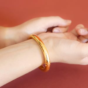 May include: A polished gold-tone hinged bangle bracelet with a smooth, rounded surface. The bracelet features a secure clasp mechanism visible on the side, encircling the wrist with a continuous, reflective metallic finish and a simple, clean design.