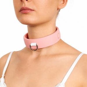 Könnte beinhalten: Ein hellrosa Halsband mit einem silberfarbenen Metallring. Das Halsband aus glattem Material ist zum Tragen um den Hals konzipiert. Es ist ein Choker-Stil.