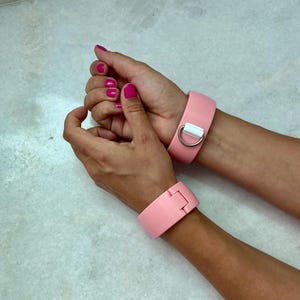 Könnte beinhalten: Zwei rosa Handfesseln mit silberfarbener Metallhardware. Eine Manschette hat einen D-Ring und einen weißen Einsatz, die andere einen Verschluss. Die Manschetten befinden sich auf einer hellen Oberfläche.