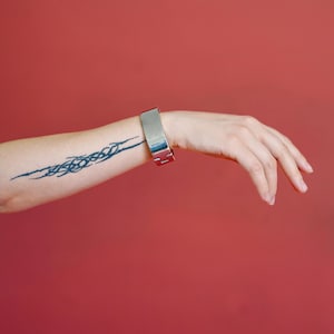 Peut inclure: Un bracelet en métal argenté de forme rectangulaire est porté au poignet. Le bracelet est le point central sur un fond rouge uni. Un tatouage bleu foncé est visible sur l'avant-bras.
