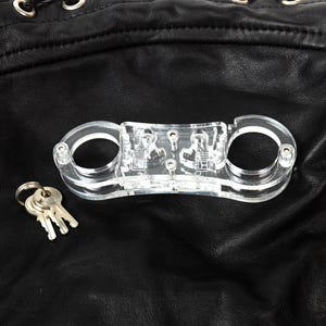 Può includere: Manette a mano in plastica trasparente con una chiave su una superficie in pelle nera.