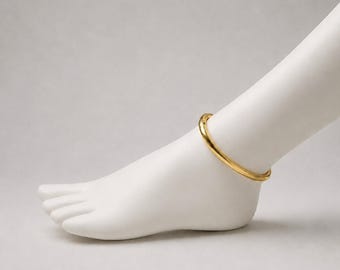 May include: A polished gold-tone anklet featuring a smooth, circular band design. The jewellery piece is displayed on a white mannequin foot, showcasing its simple, continuous metallic structure and reflective surface finish in a clear, neutral presentation.