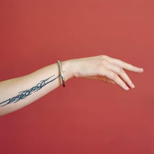 Op de afbeelding: Een zilverkleurige armband om de pols van een persoon. De armband heeft een eenvoudig, afgerond ontwerp. De arm van de persoon heeft een donkerblauwe tatoeage. De achtergrond is effen rood.