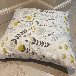 Puede incluir: Coj&iacute;n decorativo cuadrado con un estampado floral en amarillo, gris y blanco sobre fondo beige. El coj&iacute;n tiene un borde azul claro y est&aacute; sobre un suelo embaldosado.