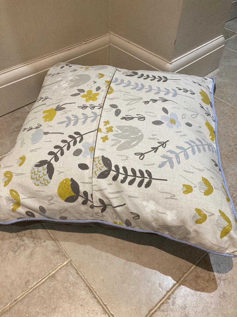 Puede incluir: Coj&iacute;n decorativo cuadrado con un estampado floral en amarillo, gris y blanco sobre fondo beige. El coj&iacute;n tiene un borde azul claro y est&aacute; sobre un suelo embaldosado.