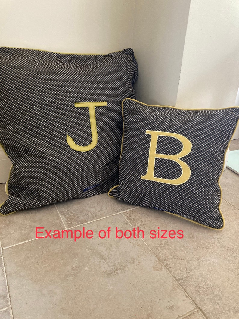 Puede incluir: Dos cojines decorativos, uno grande y otro peque&ntilde;o, ambos con un estampado de puntos azul marino y blanco. El coj&iacute;n m&aacute;s grande tiene una letra dorada "J", y el m&aacute;s peque&ntilde;o tiene una letra dorada "B". Ambos cojines tienen ribetes dorados.