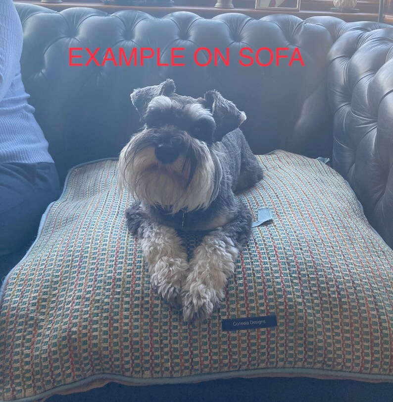 Puede incluir: Un perro peque&ntilde;o, gris y blanco descansa sobre una cama para perros estampada. La cama tiene un dise&ntilde;o tejido en tonos de azul, naranja y beige. La cama para perros tiene una etiqueta que dice "Conleis Designs". El texto "EXAMPLE ON SOFA" es visible en rojo.