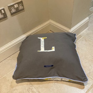 Puede incluir: Un coj&iacute;n cuadrado gris con ribete azul claro. El coj&iacute;n presenta una gran "L" aplicada en un estampado floral. El texto "Connie Designs" est&aacute; en una peque&ntilde;a etiqueta en el coj&iacute;n.