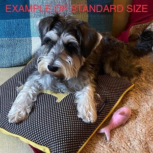 Puede incluir: Un perro peque&ntilde;o, gris y negro descansa sobre una cama para perros estampada con un borde amarillo. Un juguete rosa con forma de pez est&aacute; cerca. La cama tiene un patr&oacute;n de cuadros blancos y negros con un acento amarillo.