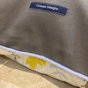 Puede incluir: Primer plano de un coj&iacute;n gris con un borde estampado floral. El borde presenta flores amarillas y grises sobre un fondo crema. Una etiqueta azul marino con el texto "Coneela Designs" est&aacute; cosida en el coj&iacute;n.