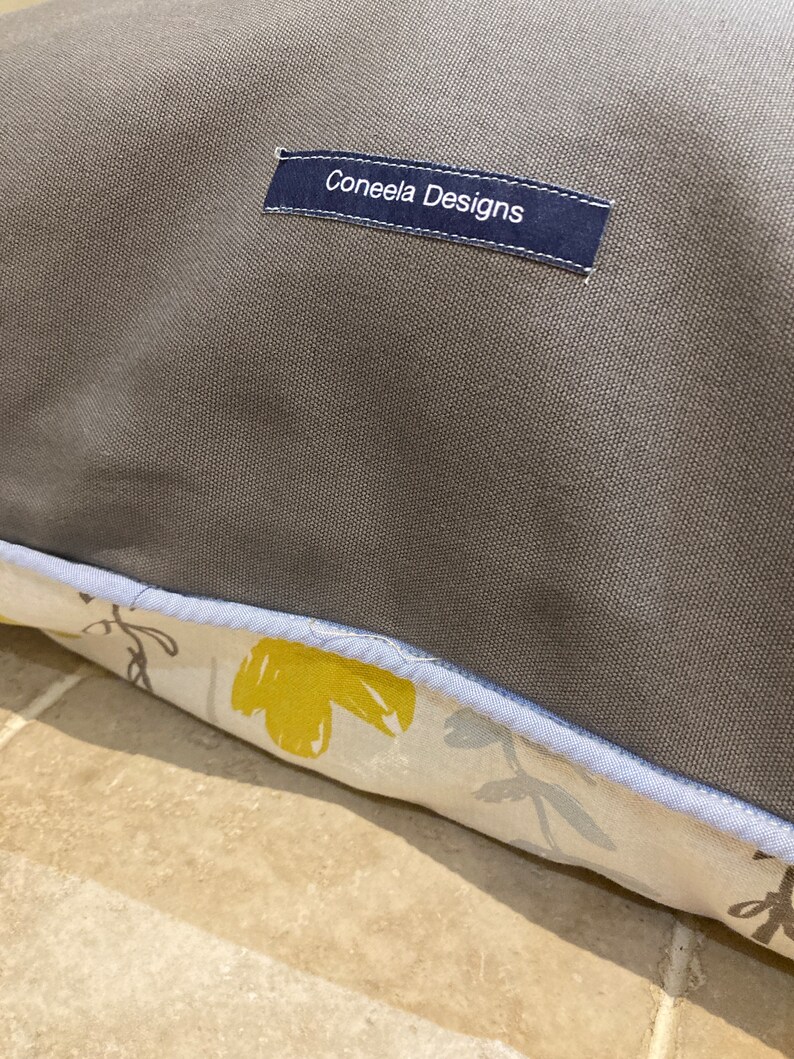 Puede incluir: Primer plano de un coj&iacute;n gris con un borde estampado floral. El borde presenta flores amarillas y grises sobre un fondo crema. Una etiqueta azul marino con el texto "Coneela Designs" est&aacute; cosida en el coj&iacute;n.