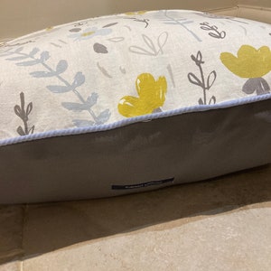 Puede incluir: Una cama rectangular para mascotas con un estampado floral. La parte superior presenta una tela color crema con flores y hojas amarillas y grises. La parte inferior es de tela gris lisa. Un ribete azul claro bordea la parte superior. Una peque&ntilde;a etiqueta con texto est&aacute; en la parte inferior.