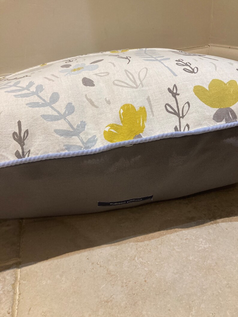 Puede incluir: Una cama rectangular para mascotas con un estampado floral. La parte superior presenta una tela color crema con flores y hojas amarillas y grises. La parte inferior es de tela gris lisa. Un ribete azul claro bordea la parte superior. Una peque&ntilde;a etiqueta con texto est&aacute; en la parte inferior.