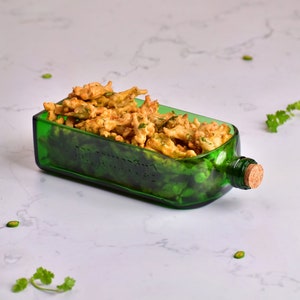 Op de afbeelding: Een hergebruikte groene glazen fles omgevormd tot een serveerschaal, gevuld met goudbruin gebakken voedsel. De fles behoudt zijn oorspronkelijke vorm, met een kurk. Groene kruiden en plakjes chilipeper voegen kleur toe.