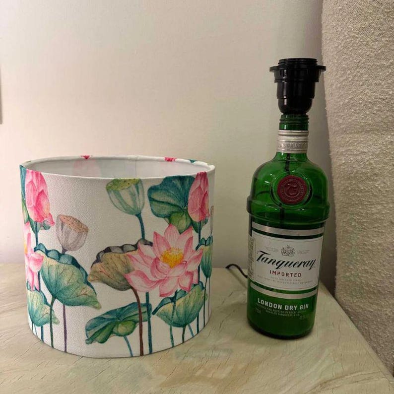 Puede incluir: Una pantalla de l&aacute;mpara blanca con un patr&oacute;n floral de nen&uacute;fares rosas y verdes. La pantalla de la l&aacute;mpara est&aacute; sobre una mesa junto a una botella de vidrio verde con una pantalla de l&aacute;mpara negra en la parte superior. La botella tiene una etiqueta que dice "Tanqueray Imported London Dry Gin".