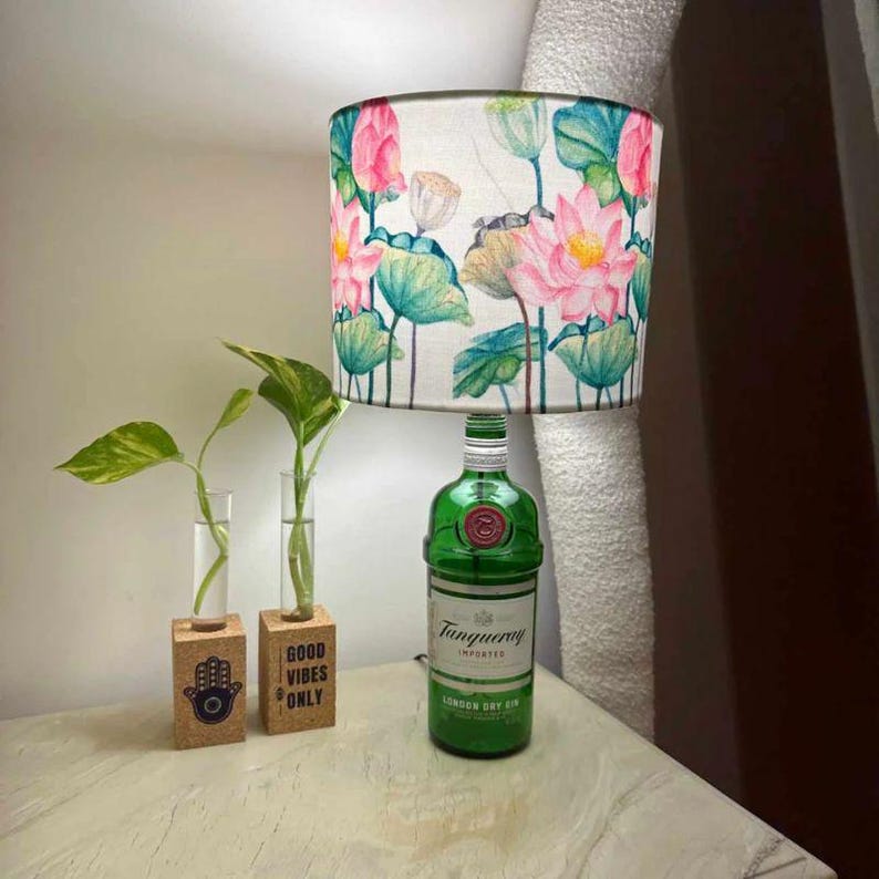 Puede incluir: Una botella de vidrio verde con una pantalla de l&aacute;mpara blanca con un dise&ntilde;o floral de flores de loto rosas y hojas verdes. La botella est&aacute; etiquetada con "Tanqueray London Dry Gin". La l&aacute;mpara est&aacute; sobre una mesa de madera con dos peque&ntilde;os jarrones de corcho con plantas.