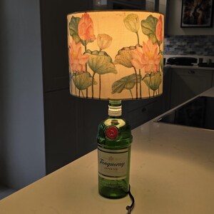 Puede incluir: Una botella de vidrio verde con una etiqueta blanca que dice "Tanqueray Importado" se utiliza como base de l&aacute;mpara. La pantalla de la l&aacute;mpara es blanca con un patr&oacute;n floral de flores de loto rosas y hojas verdes.
