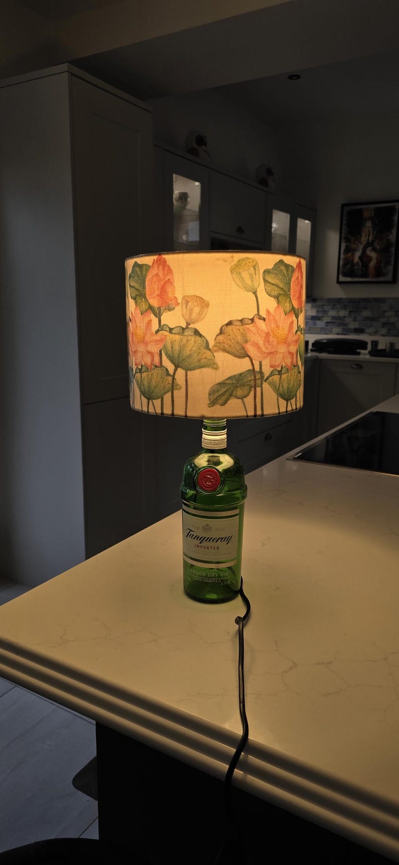 Puede incluir: Una botella de vidrio verde con una etiqueta blanca que dice "Tanqueray Importado" se utiliza como base de l&aacute;mpara. La pantalla de la l&aacute;mpara es blanca con un patr&oacute;n floral de flores de loto rosas y hojas verdes.