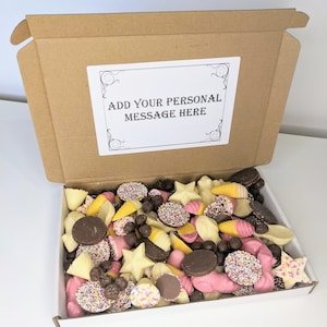 May include: A cardboard box filled with a selection of chocolate sweets. The box is open and the sweets are visible. The sweets are in a variety of shapes and colours, including white, pink, and brown. The box has a white label on the top that says "Add your personal message here".