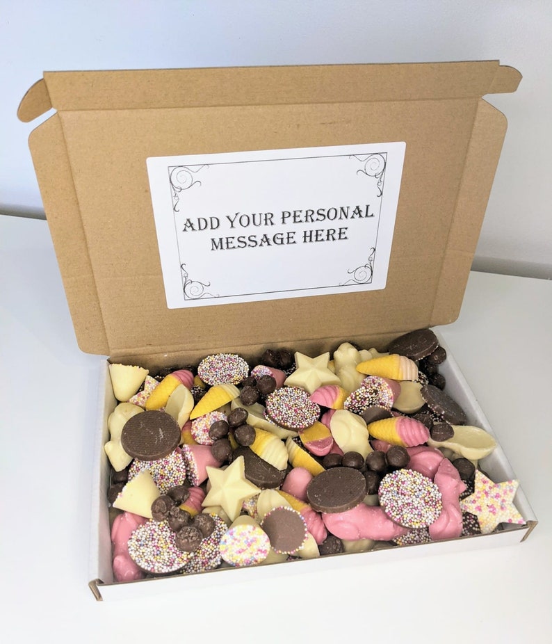 May include: A cardboard box filled with a selection of chocolate sweets. The box is open and the sweets are visible. The sweets are in a variety of shapes and colours, including white, pink, and brown. The box has a white label on the top that says "Add your personal message here".