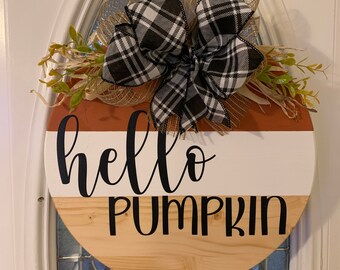 May include: A round wooden door hanger with a brown, white, and orange striped design. The words "hello pumpkin" are written in black on the hanger. The hanger is decorated with a black and white plaid bow and burlap.