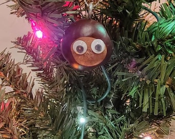 May include: A brown chestnut with googly eyes is hanging on a Christmas tree with gold and green tinsel.