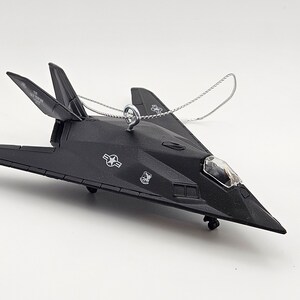 May include: Black plastic ornament of a stealth fighter jet with a silver string for hanging. The jet has a clear plastic canopy and a star on the side.