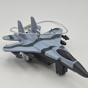 May include: A gray and black toy airplane with a silver hanging loop. The model aircraft has a black cockpit and wings, with the text "US" visible. The toy plane is a decorative ornament.