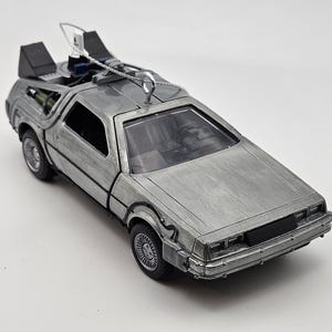May include: Silver die-cast model car with black accents, featuring a detailed exterior and interior. The car has a distinctive design with gull-wing doors and a rear-mounted engine. The model includes a silver metal loop for hanging.