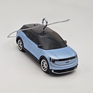 May include: A light blue Ford car ornament with a black roof and silver trim. The car has a silver loop for hanging.