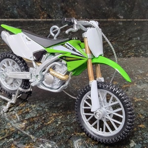 May include: A miniature Kawasaki KX 250F dirt bike model. The bike features a green and white color scheme with black tires and silver accents. The model includes a stand for display.