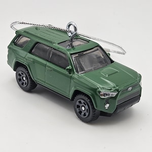 May include: A miniature green Toyota SUV model vehicle featuring black wheels, a dark interior, and a silver metal eye hook attached to the roof for hanging. The exterior paint is a glossy forest green with a small Toyota emblem on the front grille.
