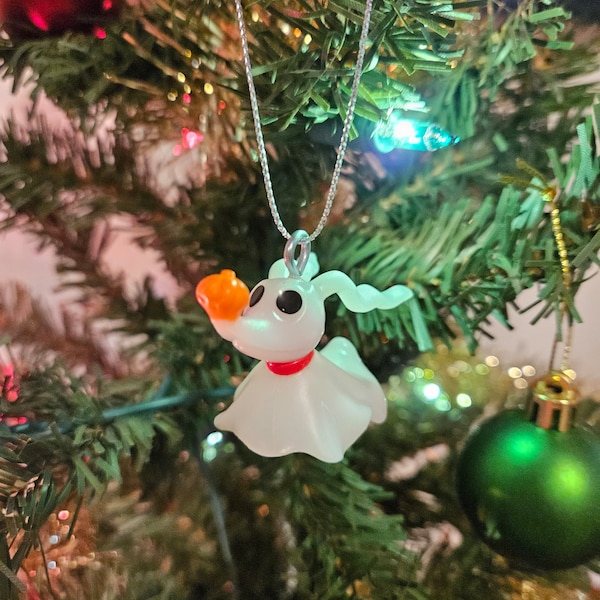 May include: A white glass ornament shaped like Zero, the ghost dog from Nightmare Before Christmas, hanging on a Christmas tree. Zero has a red collar and orange nose.