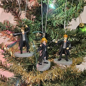 May include: Three Harry Potter character ornaments hanging on a Christmas tree. Each ornament features a figure in a black robe, standing on a gray base, with silver hanging strings. The tree is decorated with gold tinsel and green and red ornaments.
