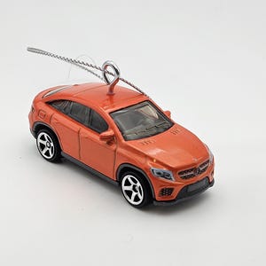 May include: Orange car ornament with a silver string for hanging. The car is a Mercedes-Benz GLE Coupe.