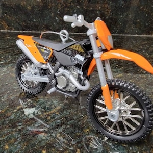 May include: A miniature orange and black KTM dirt bike ornament. The toy motorcycle features a silver exhaust pipe, wheels, and suspension. The front fender and fairing are bright orange. The tires have a knobby tread pattern. The bike has a small loop for hanging.