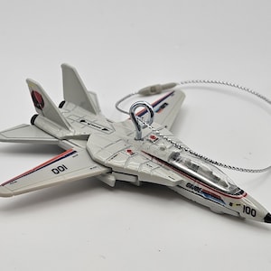 May include: A white toy airplane with red and blue accents, featuring the text "G.I. JOE" and the numbers "001" and "100". The toy has a silver string for hanging.