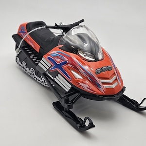 May include: A red and black toy snowmobile with a blue and white graphic design. The snowmobile has a clear windshield and a black track. The snowmobile is labeled "Turbo" and "300".