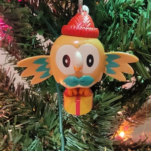 May include: A yellow and brown owl Christmas ornament with a red Santa hat, a blue mustache, and a yellow gift box. The ornament is hanging on a green Christmas tree.