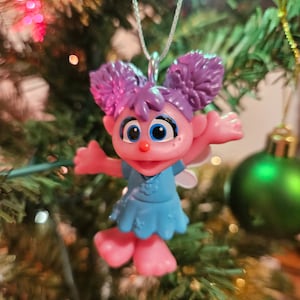 May include: A pink and purple Christmas bauble featuring a character with blue eyes, a blue dress, and purple hair. The bauble is hanging from a silver string on a green Christmas tree.